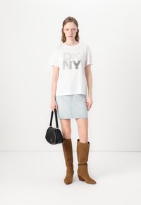 Balta marškinėliai su metaliniu "DKNY" grafiniu motyvu, mėlyna raštuota sijonas, juoda rankinė ir rudieji odiniai ilgaauliai batai ant vientiso fono.