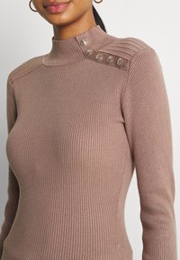 Ribbed turtleneck neulepaita vaaleanruskeana, jossa on olkapäillä napit; siinä on tiukka muotoilu ja teksturoitu kangas.