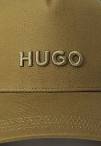 Căciulă de baseball khaki cu broderia "HUGO" în aur. Prezintă un material texturat și o margine curbată, cu detalii de cusătură vizibile.