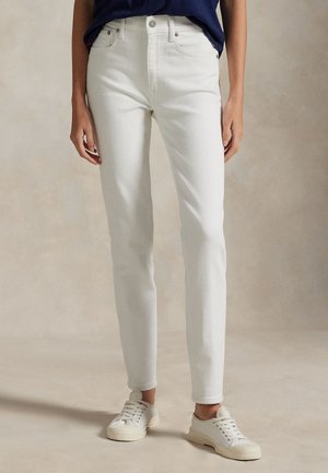 Person som bär vita smala jeans, vita sneakers och en instoppad marinblå topp mot en neutral beige bakgrund.