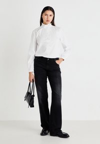 White, long-sleeve, ruffled blouse with button closure; paired with black, slightly flared jeans; accessorized with a small black handbag.