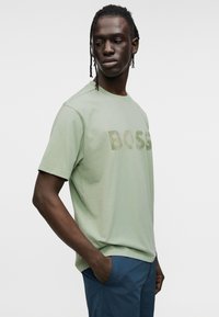 Ανοιχτό πράσινο βαμβακερό t-shirt με τη λέξη "BOSS" τυπωμένη σε υφή, ελαφρώς πιο σκούρο χρώμα. Λαιμόκοψη τύπου crew και κοντά μανίκια.