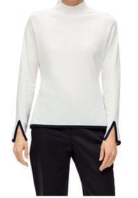 Vit långärmad topp med svartkantade slits och nederkant, kombinerad med svarta byxor på en person mot en vit bakgrund.