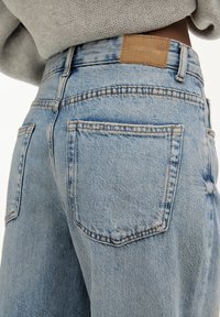 Jeans in denim azzurro chiaro con vestibilità rilassata, dotati di tasche posteriori, cuciture in evidenza e un'etichetta in pelle marrone sulla cintura.