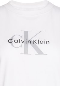 Λευκό πουκάμισο με το λογότυπο Calvin Klein, με μεγάλα ξεθωριασμένα γράμματα "CK" στο φόντο και "Calvin Klein" κεντημένα μαύρα στο στήθος.