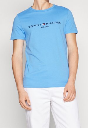 T-shirt en coton bleu avec col rond, manches courtes, affichant le texte « TOMMY HILFIGER EST. 1985 » en noir et un petit détail de logo sur la manche.