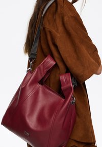 Raudona odinė rankinė su išpjautomis rankenomis ir pilku reguliuojamu diržu, pasižyminti lygiu paviršiumi ir minimalistiniu dizainu. Sidabro spalvos aksesuarai.