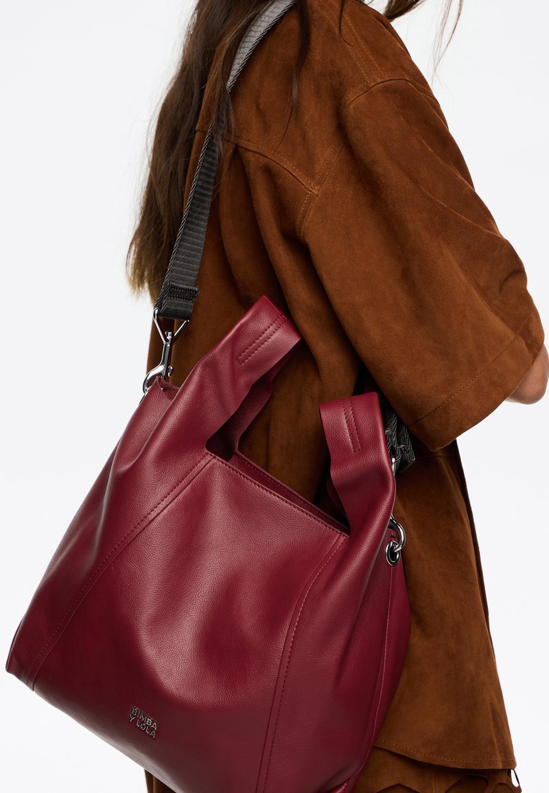 Raudona odinė rankinė su išpjautomis rankenomis ir pilku reguliuojamu diržu, pasižyminti lygiu paviršiumi ir minimalistiniu dizainu. Sidabro spalvos aksesuarai.