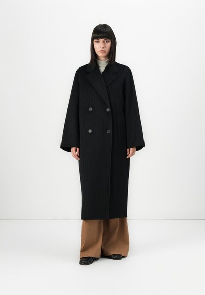 Donna in piedi con capelli neri lunghi e lisci, che indossa un lungo cappotto doppiopetto a righe nere, pantaloni beige a gamba larga e scarpe nere.