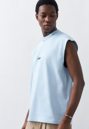 Νεαρός άνδρας με ανοιχτόμπλε αμάνικο πουκάμισο και μπεζ παντελόνι κοιτάζει προς τα κάτω, φοράει ασημένιο βραχιόλι και κολιέ, σε απλό φόντο.