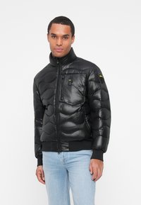 Giacca a vento nera con alto colletto, texture trapuntata, chiusura frontale con zip e taschino sul petto. Presenta una finitura morbida e lucida.