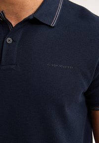 Námořnická modrá polo košile vyrobená z texturované tkaniny; obsahuje límec se pruhovanými akcenty a zapínání na knoflíky; logo vyšívané na hrudi.