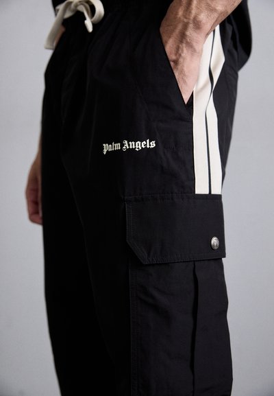 Man wearing black cargo pants with white side stripes and "Palm Angels" logo, hand in pocket, against a neutral background.