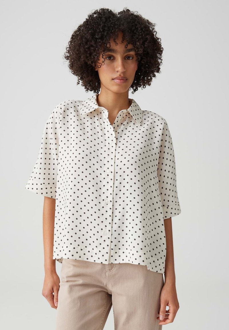 Young woman with curly hair wearing a white short-sleeve button-up shirt with black polka dots and light beige pants.