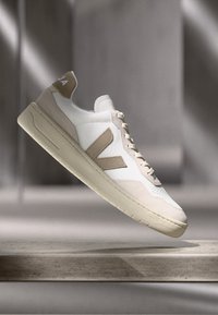 Hvite sneakers med beige detaljer, med en glatt læroverdel, strukturerte sider, flate lisser og en gummisåle med et minimalistisk design.