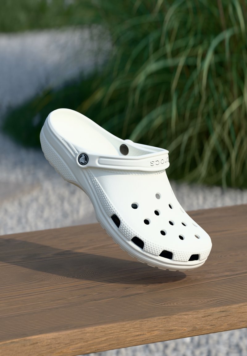 Sabot blanc sans lacets avec trous de ventilation et bride au talon, posé sur une surface en bois à l'extérieur avec de la verdure en arrière-plan.