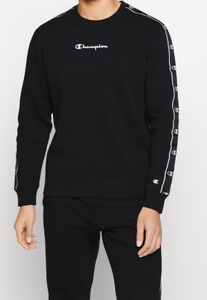 Mand iført sort Champion sweatshirt og matchende bukser, sweatpants har logo-striber ned ad ærmerne og buksebenene.