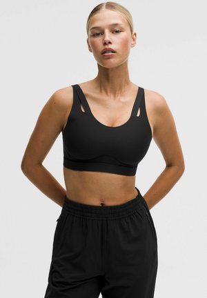 Ung kvinde står med hænderne bag ryggen iført en sort sports-bh og sorte sportbukser mod en ensfarvet lys baggrund.