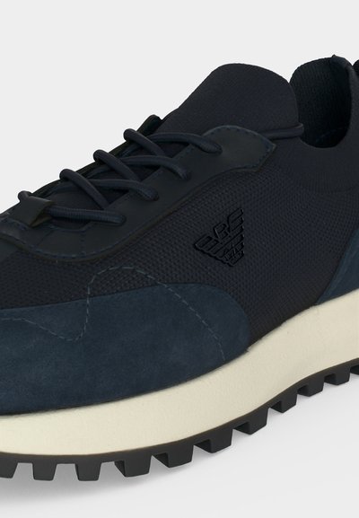 Gros plan d'une basket bleu foncé en tissu texturé, avec des accents en daim, des lacets noirs, une semelle intermédiaire blanche et une semelle extérieure noire à crampons.