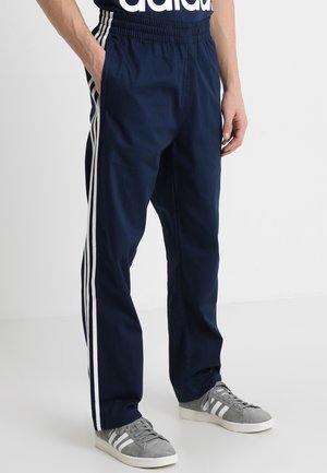 Hombre con pantalones deportivos Adidas azul marino con rayas blancas a los lados y zapatillas Adidas grises, de pie con las manos en los bolsillos.