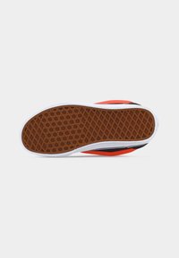 Semelle de chaussure avec motif gaufré marron en forme de gaufre, bord de semelle intermédiaire blanc, et parties supérieures visibles rouges et noires sur un fond gris clair.
