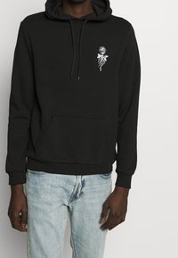 Svart hoodie med framficka i form av en känguru, dragsko i huvan och vitt ros-motiv på vänster bröst. Tillverkad av mjukt tyg med ribbade ärmslut.