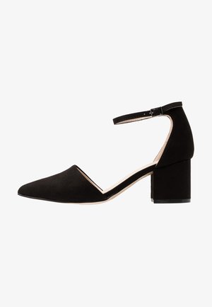 Sapato preto em camurça com salto bloco, bico fino e faixa no tornozelo; apresenta uma textura suave e um design minimalista.