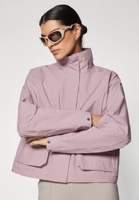 Light pink waterproof jacket with a high collar, two front pockets, and snap-button closures. Smooth texture, fitted design, minimalistic style.