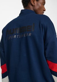 Giacca sportiva blu navy con colletto alto, caratterizzata da un logo nero sulla schiena e accenti in rosso e bianco a blocchi di colore sulle maniche.