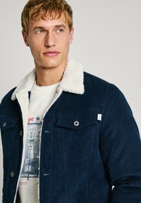 Jachetă din corduroy navy cu guler din blană artificială crem, având două buzunare pe piept și nasturi metalici pentru închidere. Textură ușoară.