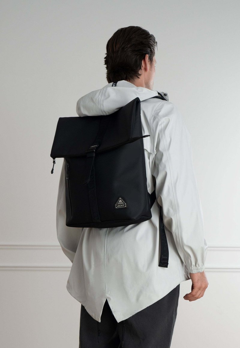 Mochila negra con tapa plegable, hecha de material duradero. Cuenta con un bolsillo frontal con cremallera y un logotipo en forma de triángulo. Se lleva en la espalda.