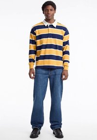 Dlhé rukávy polo tričko so žltomodrými pruhmi a bielym golierom, kombinované s modrými džínsami a čiernymi topánkami. Má voľný strih.