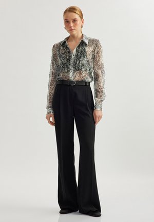 Kvinde står iført en gennemsigtig bluse med dyreprint og volanger, sorte højtaljede bukser med brede ben, sort bælte og sorte sko foran en hvid baggrund.