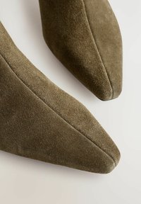 Tacones de ante verde oliva con punta afilada, de textura suave y costuras limpias, que presentan una silueta elegante y un diseño minimalista.