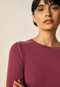 Mauve színű, hosszú ujjú felső bordás textúrával, kerek nyakkivágással és látható vállvarrásokkal. A modellen keresztbe tett karokkal, kis karikafülbevalóval látható.
