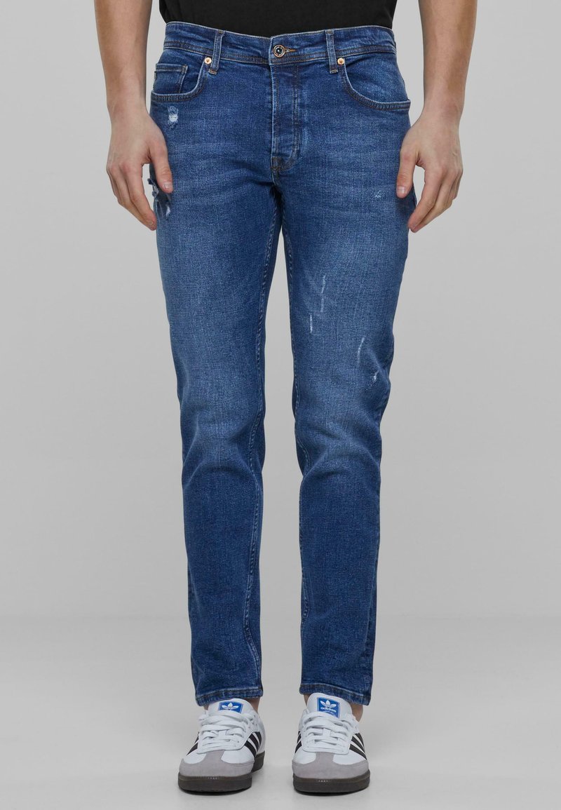 Jeans de mezclilla azul con un corte ajustado, que presentan acentos desgastados y un ligero desgarro. Combinados con zapatillas blancas y grises con rayas negras.