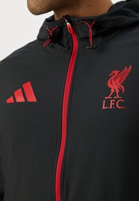 Sort træningsjakke med rød lynlås og detaljer. Har L.F.C. logo og tre røde striber på skulderen. Teksturdesign i sporty stil.