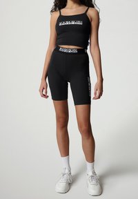 Reggiseno sportivo nero e pantaloni corti abbinati con dettagli del logo sul davanti. Tessuto liscio; design aderente. Scarpe e calze da ginnastica bianche.