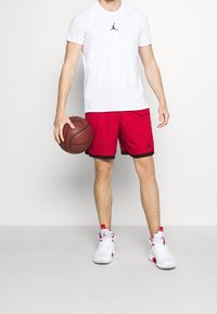 Hombre con camiseta blanca de Jordan, pantalones cortos de baloncesto rojos y zapatillas blanco-rojas, sosteniendo un balón de baloncesto con su mano izquierda frente a un fondo blanco.