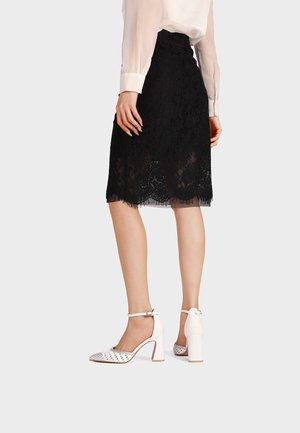 Mujer vestida con una falda negra de encaje hasta la rodilla, blusa blanca transparente y zapatos blancos de tacón alto con correas en el tobillo, de pie frente a un fondo simple.