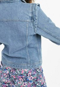 Světle modrá džínová bunda s klasickým límcem, manžetami na knoflíky a viditelným prošitím. Natažená přes květinový vzor na spodní části.