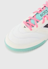 Hvit sneaker med rosa lisser og en teal mesh tunge. Har en gjennomsiktig mintgrønn såle og en teksturert hvit overdel med svarte detaljer.