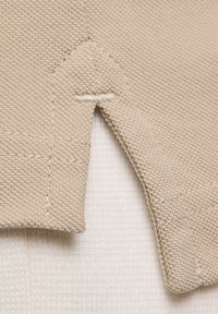 Tessuto a maglia beige con una superficie texturizzata. Presenta una cucitura visibile e una sezione attaccata, con una finitura pulita lungo i bordi.