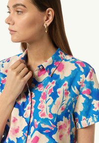 Camicia a maniche corte floreale in crema con motivi blu e rosa. Presenta un colletto con bottoni e piccoli bottoni lungo la parte anteriore.