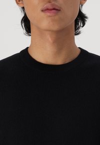 Nahaufnahme einer Person, die einen schwarzen, gerippten Rundhalsausschnitt-Pullover trägt, wobei unteres Gesicht, Hals und Schultern vor einem schlichten Hintergrund zu sehen sind.