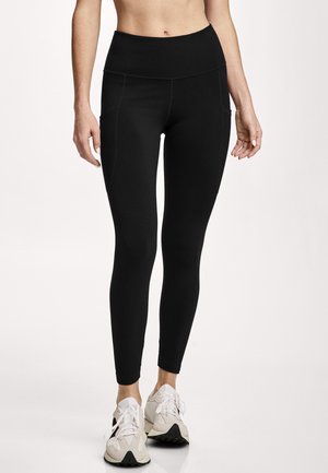 Woman wearing high-waisted black leggings with side pockets and white athletic shoes, standing against a plain light background.