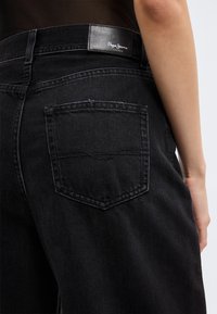 Μαύρα τζιν από τζιν με πίσω τσέπη και ετικέτα Pepe Jeans London, που φοριούνται από άτομο με γυμνό χέρι ορατό στη δεξιά πλευρά.