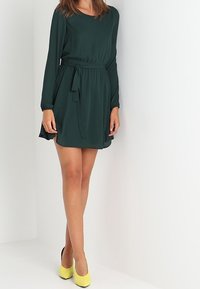 Grünes Langarmkleid mit taillierter Taille und fließendem Rock. Verfügt über einen Rundhalsausschnitt und eine strukturierte Stoffbeschaffenheit. Kombiniert mit gelben High Heels.