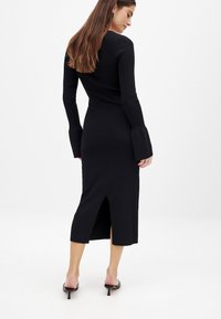 Musta ribbimittainen mekko pitkillä hihoilla ja levenevillä hihansuilla. Sisältää takaläpän ja vartalonmyötäisen siluetin, yhdistettynä matalakorkoisiin mustiin kenkiin.