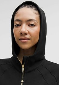 Sudadera con cremallera negra y un cuello alto, de textura de tela sustancial. El hardware dorado adorna la cremallera, creando un diseño elegante.